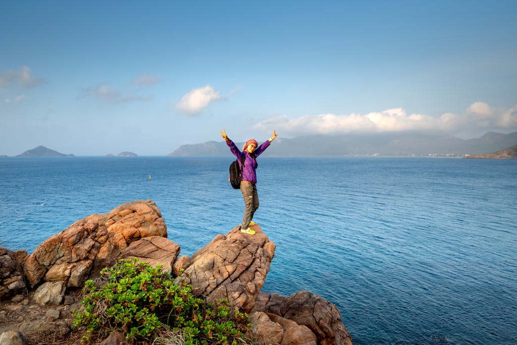 A backpacker stands triumphantly on a rocky cliff overlooking the sea, embracing outdoor adventure.