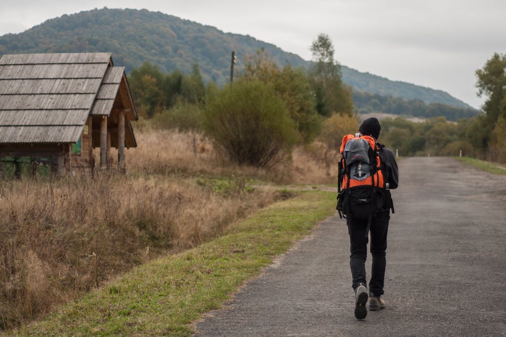 Back view full body of anonymous hiker with backpack strolling on asphalt roadway near shabby house inn suburb area during trekking